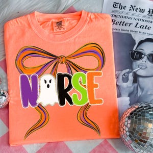 May include: A neon orange t-shirt with a graphic design featuring the word "NURSE" in purple, black, green, and orange, with a ghost in place of the "U". A large bow with purple and orange accents is above the word. The shirt is a Comfort Colors brand.
