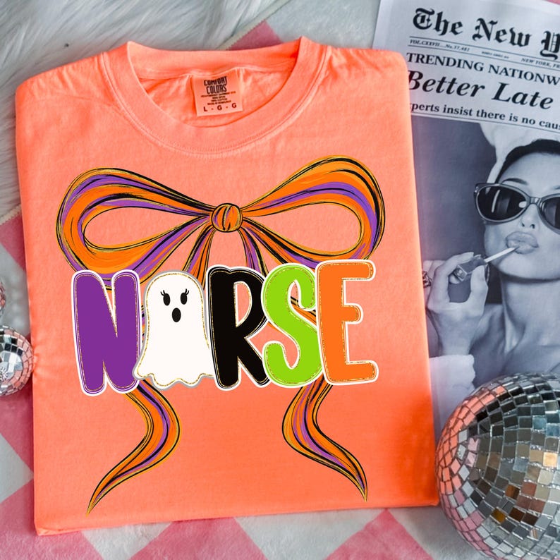 May include: A neon orange t-shirt with a graphic design featuring the word "NURSE" in purple, black, green, and orange, with a ghost in place of the "U". A large bow with purple and orange accents is above the word. The shirt is a Comfort Colors brand.