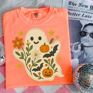 May include: Coral-coloured t-shirt featuring an embroidered ghost design with flowers, pumpkins, bats, and stars. The crew neck shirt has the brand name "Comfort Colors" on the tag. A fun, seasonal design.