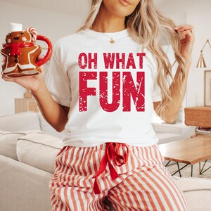 May include: White t-shirt with the red text "OH WHAT FUN". The person is wearing red and white striped pajama pants and holding a gingerbread man mug with a red handle. The mug has a red scarf and candy cane.