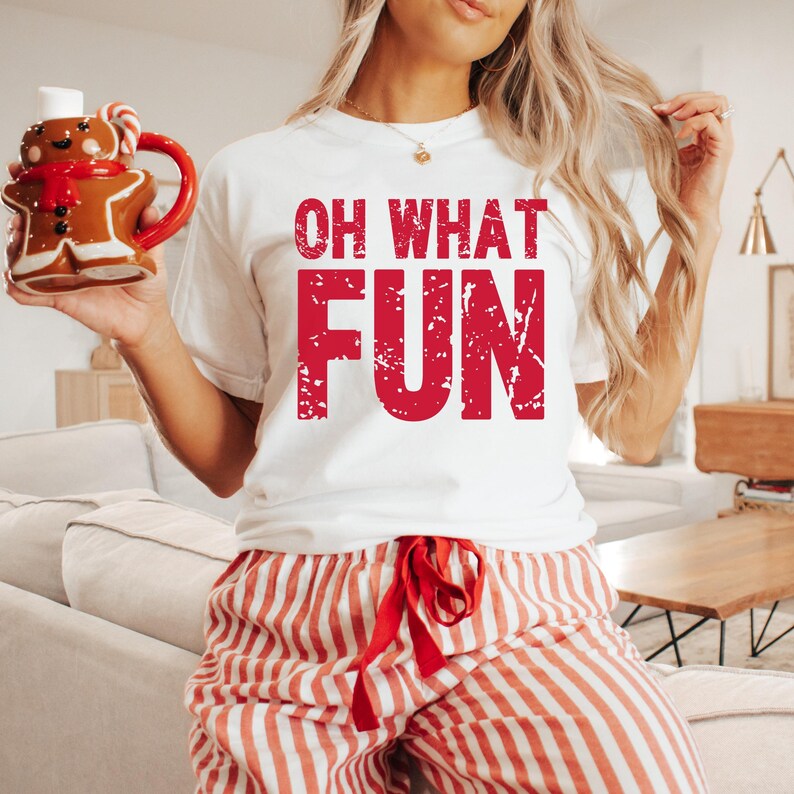 May include: White t-shirt with the red text "OH WHAT FUN". The person is wearing red and white striped pajama pants and holding a gingerbread man mug with a red handle. The mug has a red scarf and candy cane.