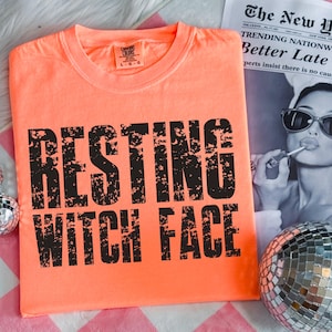 May include: An orange t-shirt with the phrase "RESTING WITCH FACE" printed in large, distressed black letters. The shirt is a Comfort Colors brand. A newspaper and a disco ball are in the background.