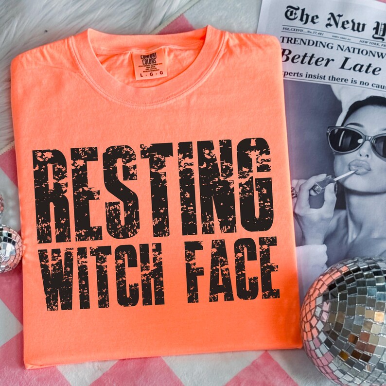 May include: An orange t-shirt with the phrase "RESTING WITCH FACE" printed in large, distressed black letters. The shirt is a Comfort Colors brand. A newspaper and a disco ball are in the background.