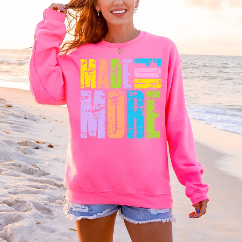 May include: A bright pink sweatshirt with the words "MADE MORE" in large, colorful, distressed text across the front. The letters are in shades of yellow, blue, green, and purple. The background is a beach scene.