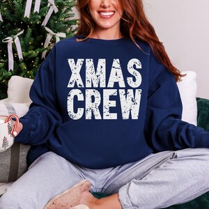 May include: Navy blue sweatshirt with the text "XMAS CREW" in distressed white lettering. The sweatshirt is a crew neck style and is being worn by a person. The background includes a Christmas tree and a mug.