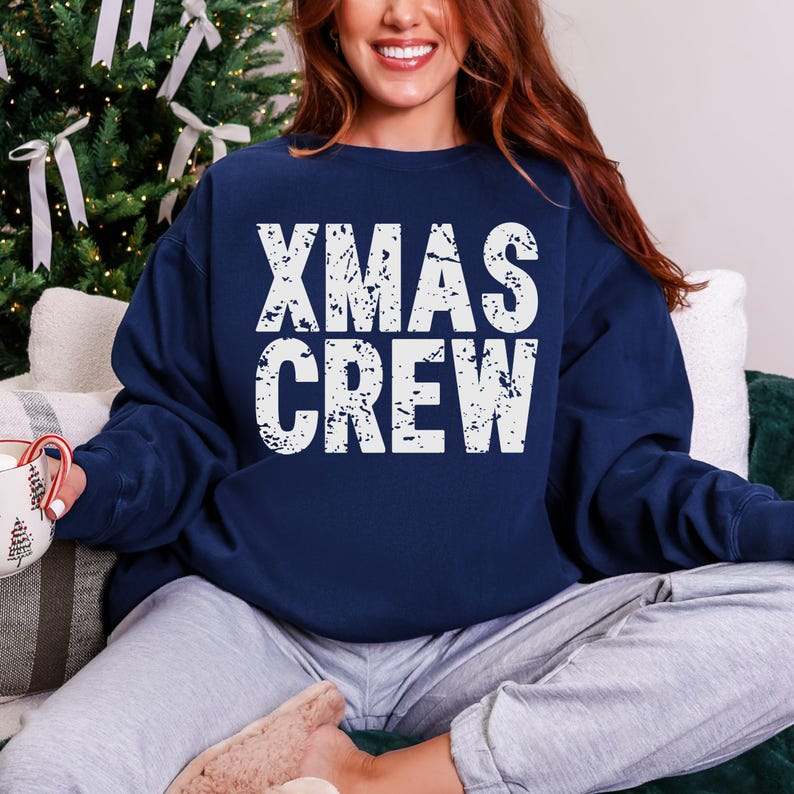 May include: Navy blue sweatshirt with the text "XMAS CREW" in distressed white lettering. The sweatshirt is a crew neck style and is being worn by a person. The background includes a Christmas tree and a mug.
