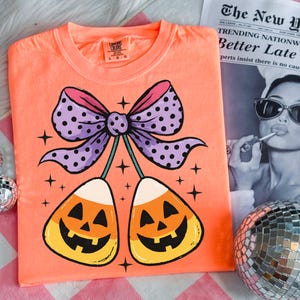 May include: Orange Comfort Colors t-shirt featuring a Halloween-themed design. The graphic includes two candy corn pieces with jack-o'-lantern faces, connected by a purple bow with black polka dots. The shirt also has the text "COMFORT COLORS".