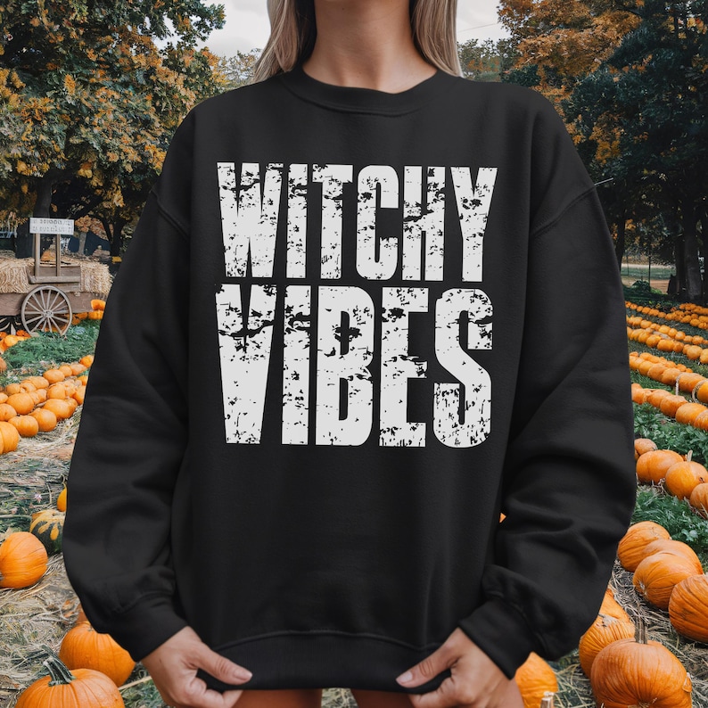 May include: Black sweatshirt with the text "Witchy Vibes" in distressed white lettering. The sweatshirt is displayed in an outdoor setting with pumpkins, suggesting a fall or Halloween theme.