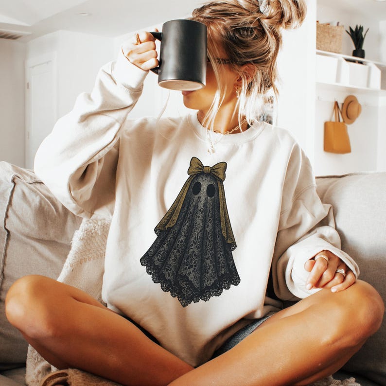 K&ouml;nnte beinhalten: Cremefarbenes Sweatshirt mit einem Geisterdesign. Der Geist hat einen schwarzen, spitzenartigen K&ouml;rper, zwei Augenl&ouml;cher und eine goldene Schleife. Das Design befindet sich auf der Vorderseite des Sweatshirts. Eine Person h&auml;lt eine schwarze Tasse.