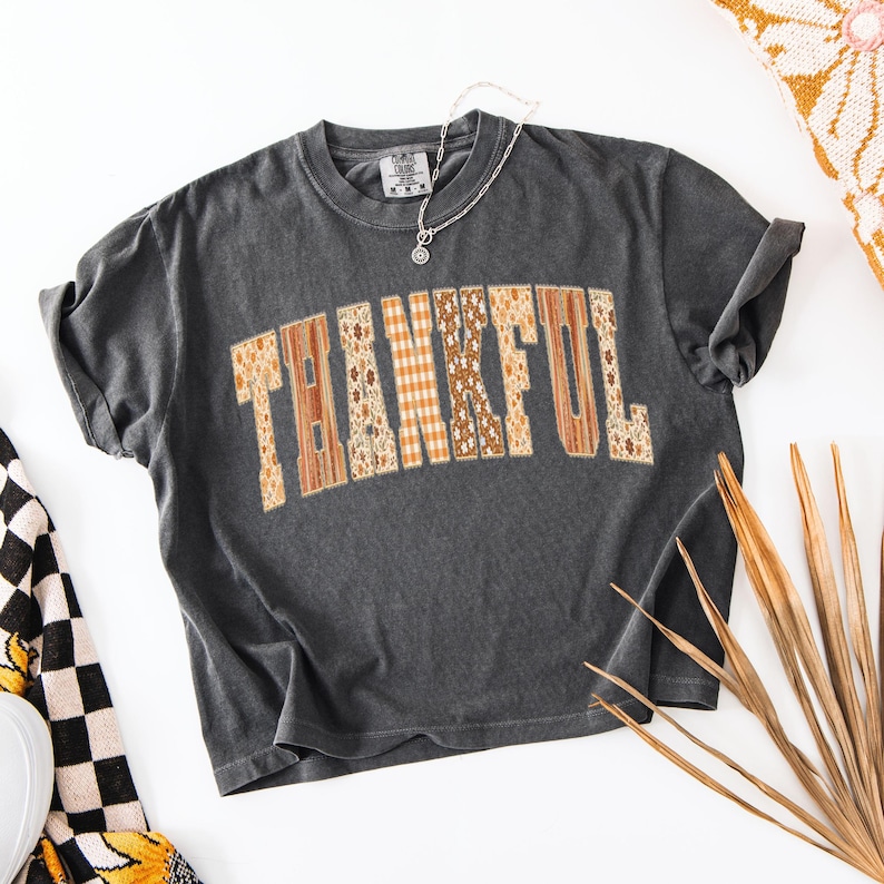 May include: A charcoal gray t-shirt with the word "THANKFUL" in a patchwork design across the front. The letters are in shades of brown, orange, and cream, with floral and plaid patterns. A silver necklace with a pendant is draped on the shirt.