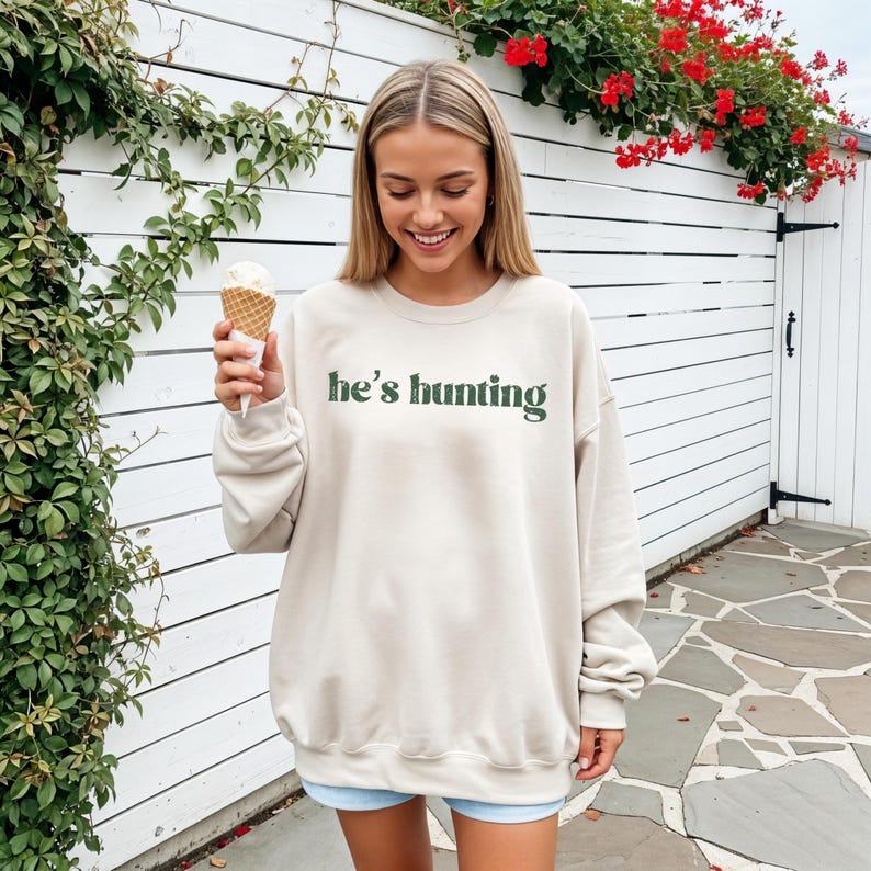 May include: A beige sweatshirt with the text "he's hunting" in green lettering. The sweatshirt is worn by a person holding an ice cream cone. The background features a white fence and greenery.
