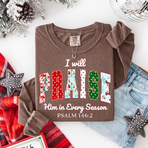 May include: A brown long-sleeve shirt with the text "I will PRAISE Him in Every Season" and Psalm 146:2. The word "PRAISE" is in a colorful, patterned font. A silver necklace and a pair of blue jeans are also visible.
