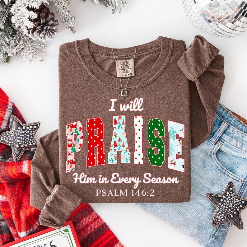 May include: A brown long-sleeve shirt with the text "I will PRAISE Him in Every Season" and Psalm 146:2. The word "PRAISE" is in a colorful, patterned font. A silver necklace and a pair of blue jeans are also visible.
