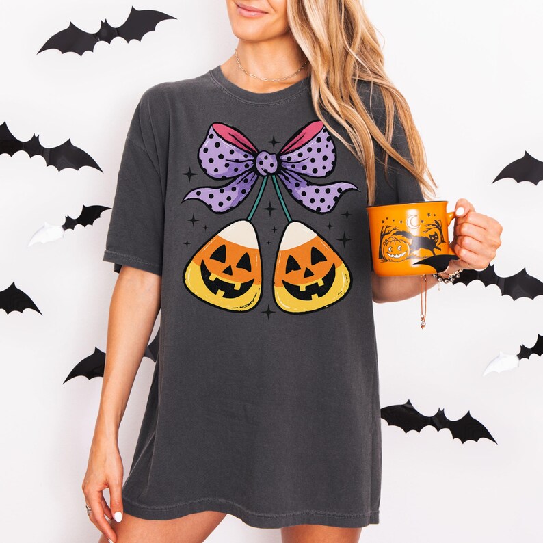 May include: Dark gray t-shirt featuring a Halloween candy corn design with a purple polka dot bow. The shirt also has two candy corn graphics with jack-o'-lantern faces. An orange mug with a Halloween design is held in the hand.