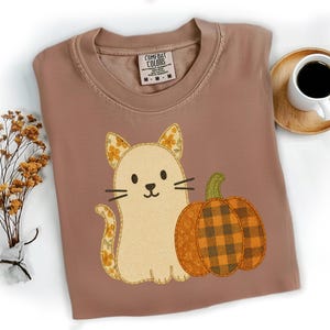 May include: A brown Comfort Colors t-shirt featuring a beige cat with floral accents and a pair of orange plaid pumpkins. The shirt has a crew neck and a tag that reads "Comfort Colors". A cup of coffee sits on a wooden coaster.
