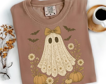 Pode incluir: Camiseta Comfort Colors marrom com um desenho de fantasma com laço, rodeado por abóboras, flores, morcegos e uma teia de aranha. O fantasma está usando um lençol rendado. Uma xícara de café em uma base de madeira.