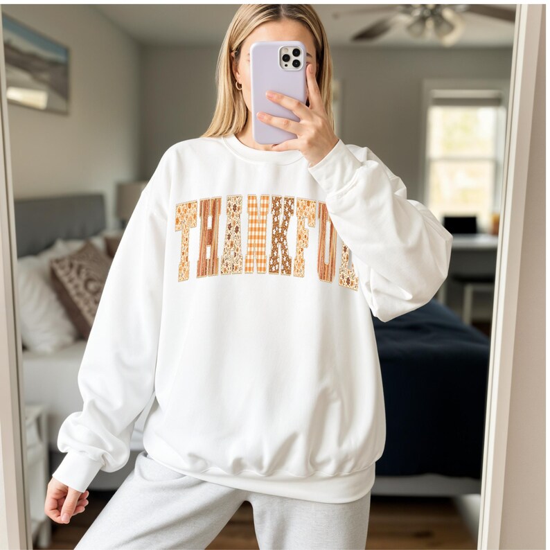 May include: White sweatshirt with the word "THANKFUL" in a patchwork design across the chest. The letters feature various fall-themed patterns in shades of brown, orange, and cream. The sweatshirt has long sleeves and a crew neck.