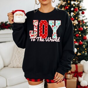 May include: Black sweatshirt with the words "JOY To The World" in a festive design. The letter "O" is red with white polka dots. The letters "J" and "Y" have a holiday pattern. A Santa mug is held in the left hand.