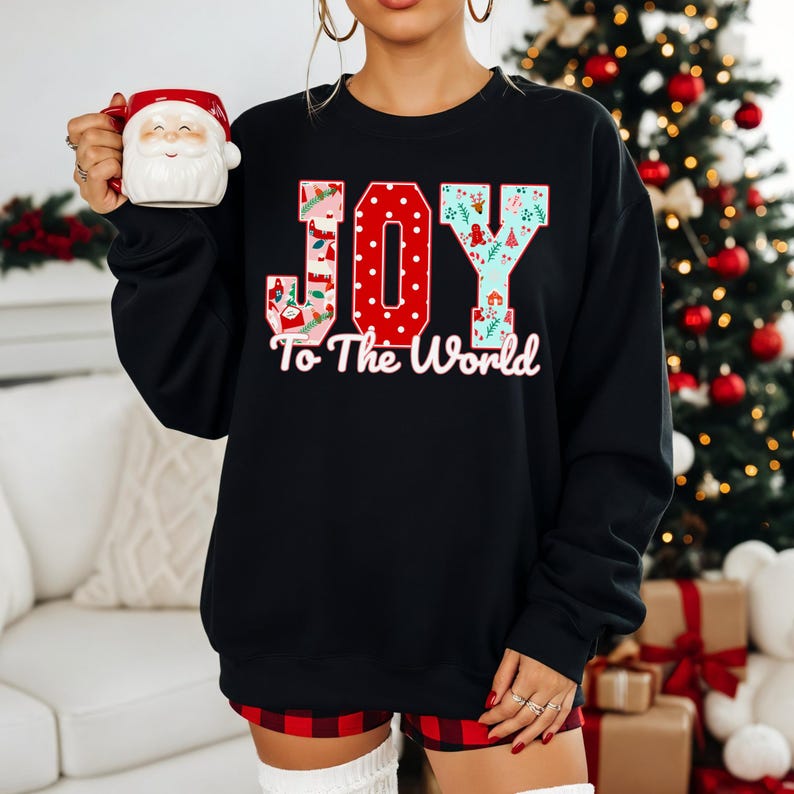 May include: Black sweatshirt with the words "JOY To The World" in a festive design. The letter "O" is red with white polka dots. The letters "J" and "Y" have a holiday pattern. A Santa mug is held in the left hand.
