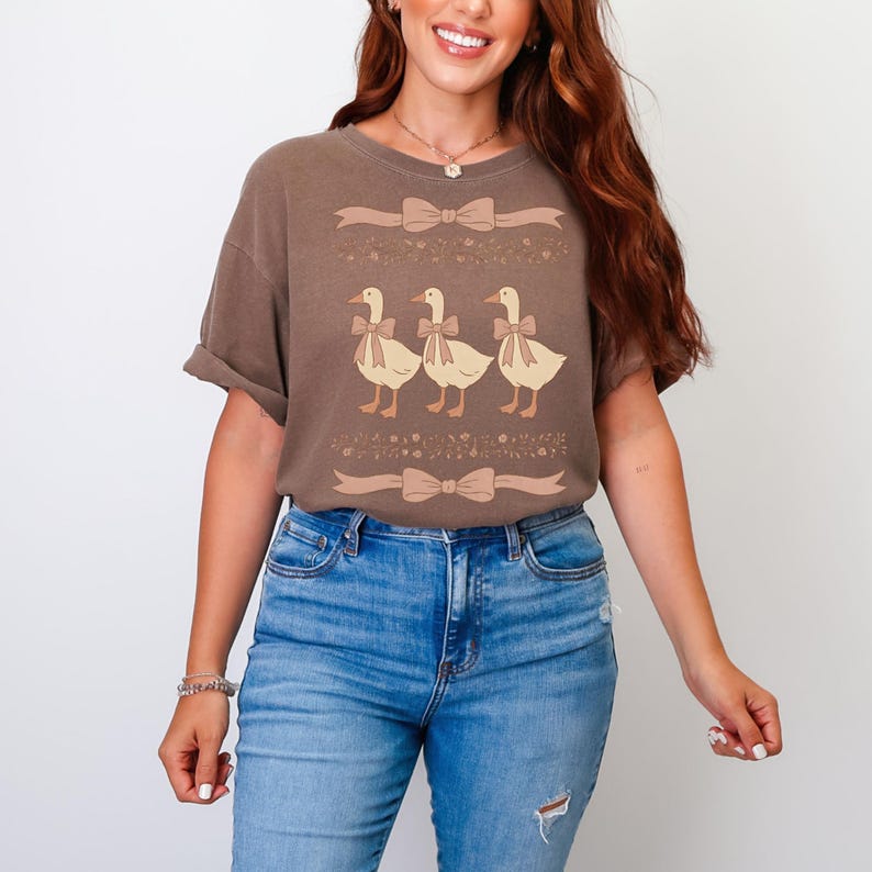 May include: A brown t-shirt with a graphic of three ducks wearing bows. The ducks are in a line and are facing the right. The shirt has a floral pattern around the ducks and a bow at the bottom.