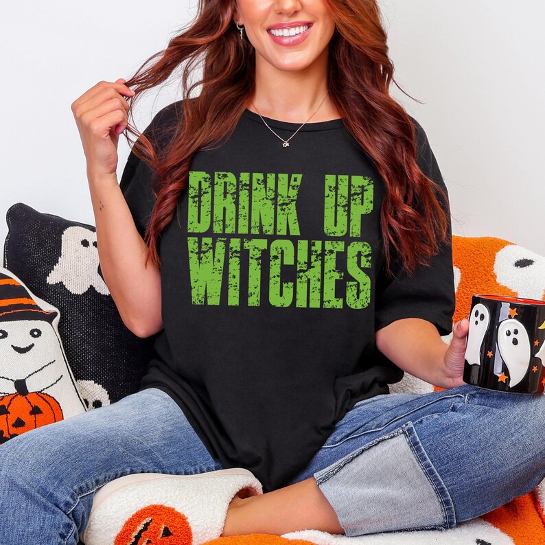 May include: Black t-shirt with the text "DRINK UP WITCHES" in distressed green lettering. The shirt is a crew neck style. The image also shows a Halloween-themed mug and decorative pillows.