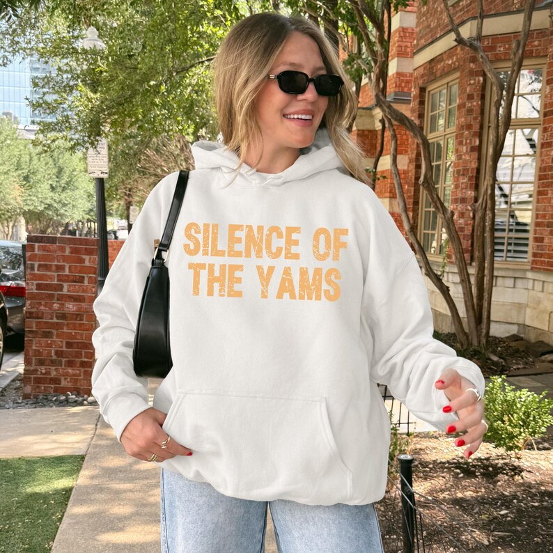 Pu&ograve; includere: Felpa con cappuccio bianca con la scritta "SILENCE OF THE YAMS" in arancione. La felpa ha una tasca frontale e un cappuccio con coulisse. La persona nell'immagine indossa occhiali da sole e jeans azzurri.