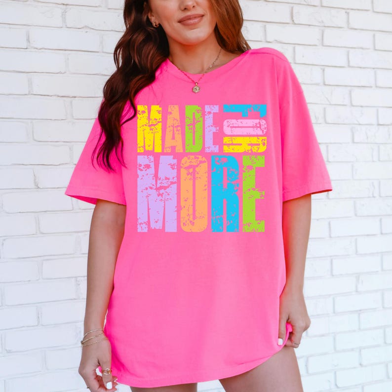 May include: A bright pink t-shirt with the words "MADE MORE" in a distressed, multi-colored font. The text is in yellow, blue, green, purple, and orange. The shirt has short sleeves and a relaxed fit.