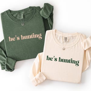 May include: Two long-sleeved shirts, one olive green and one cream, with the phrase "he's hunting" printed on the front. Each shirt has a crew neck and a small sunburst pendant necklace. The shirts are laid flat on a white surface.