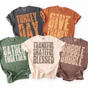 May include: Five different-colored t-shirts with Thanksgiving-themed phrases printed in distressed orange and white lettering. The shirts are in shades of gray, orange, green, cream, and brown. Phrases include "Turkey Day", "Give Thanks", "Gather Together", "Thankful Grateful Blessed", and "Gobble Gobble".