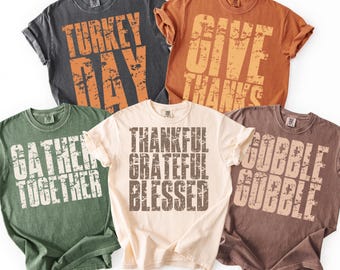 May include: Five different-colored t-shirts with Thanksgiving-themed phrases printed in distressed orange and white lettering. The shirts are in shades of gray, orange, green, cream, and brown. Phrases include "Turkey Day", "Give Thanks", "Gather Together", "Thankful Grateful Blessed", and "Gobble Gobble".