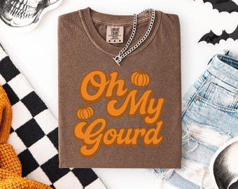 May include: A brown t-shirt with the phrase "Oh My Gourd" in orange lettering and pumpkin graphics. A silver chain necklace rests on the shirt. The shirt is surrounded by Halloween-themed decorations, including a skull and bat cutouts.