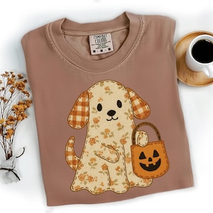 Könnte beinhalten: Ein Comfort Colors T-Shirt in Staubrosa mit einem Hund-Geist-Design mit Blumen- und Karomustern. Der Hund hält eine orangefarbene Kürbistasche. Eine Kaffeetasse steht auf einem Holzuntersetzer.