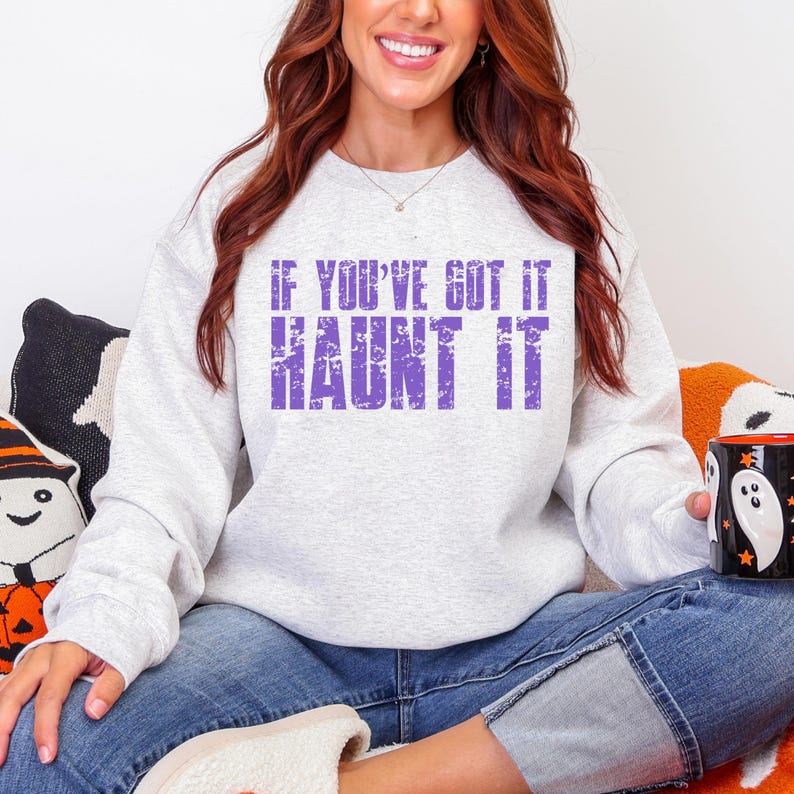 May include: A light gray sweatshirt with the text "IF YOU'VE GOT IT HAUNT IT" in purple. The sweatshirt is a crew neck style. The image is of a person wearing the sweatshirt.