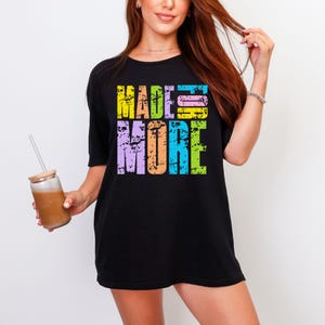May include: Black t-shirt with the words "MADE MORE" in a colorful, distressed font. The text is in yellow, pink, blue, orange, and purple. The shirt has short sleeves.