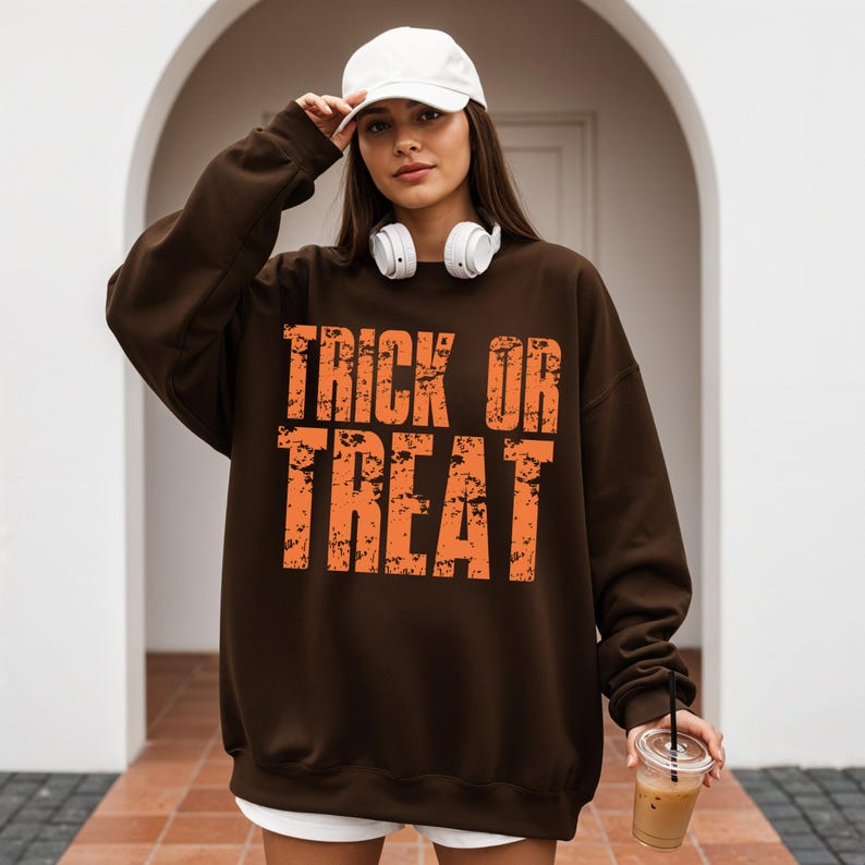 May include: A brown sweatshirt with the words "TRICK OR TREAT" in orange, distressed lettering. The sweatshirt is worn with a white baseball cap and headphones. A clear plastic cup with a straw is held in the hand.
