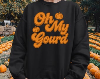 May include: Black sweatshirt with the phrase "Oh My Gourd" in orange script, featuring pumpkin illustrations. The background shows a pumpkin patch, perfect for autumn and fall-themed events.