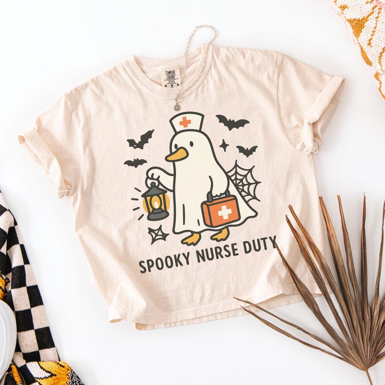 May include: Beige t-shirt with a Halloween-themed graphic. The design features a cartoon duck dressed as a nurse, holding a lantern and a medical bag. The text "SPOOKY NURSE DUTY" is printed below the graphic, with bats and a spiderweb.