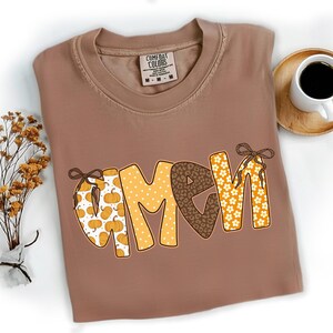 May include: A brown Comfort Colors t-shirt with the word "GMA" in a pumpkin, leopard print, and floral design. A cup of coffee sits on a wooden coaster to the right of the shirt. A small bouquet of dried flowers is to the left.