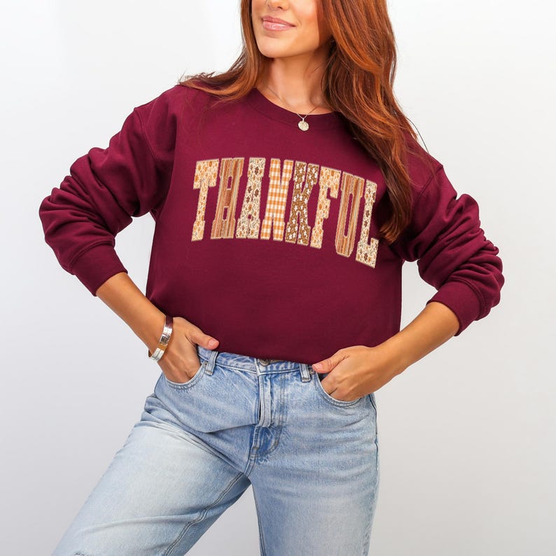 May include: Burgundy sweatshirt with the word "THANKFUL" in a patchwork design across the chest. The letters are made of various fall-themed patterns. The sweatshirt has long sleeves and a crew neck.