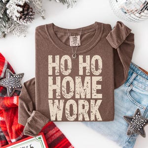 May include: A brown long-sleeve shirt with the text "HO HO HOME WORK" in a distressed, off-white font. The shirt is folded, with a silver necklace and a pair of blue jeans visible. Festive holiday theme.