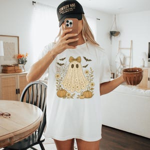 Puede incluir: Camiseta blanca con un dise&ntilde;o de fantasma vintage, con un lazo, calabazas, flores y murci&eacute;lagos. El fantasma tiene una apariencia de encaje. La camiseta es de estilo casual y c&oacute;modo.