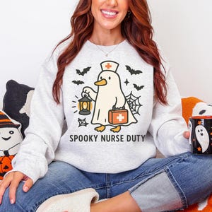 May include: A light gray sweatshirt with a Halloween-themed graphic. The design features a cartoon ghost duck wearing a nurse's hat and holding a lantern and medical bag. The text "SPOOKY NURSE DUTY" is below the graphic, with bats and a spiderweb.
