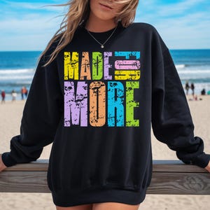 May include: Black sweatshirt with the text "MADE MORE" in a distressed, multi-colored font. The text is in yellow, pink, blue, purple, orange, and green. The sweatshirt is a casual style.