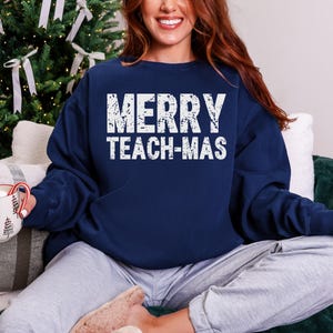 May include: Navy blue sweatshirt with the text "MERRY TEACH-MAS" in distressed white lettering. The sweatshirt is a crew neck style and is being worn by a person. The background includes a Christmas tree and a mug.