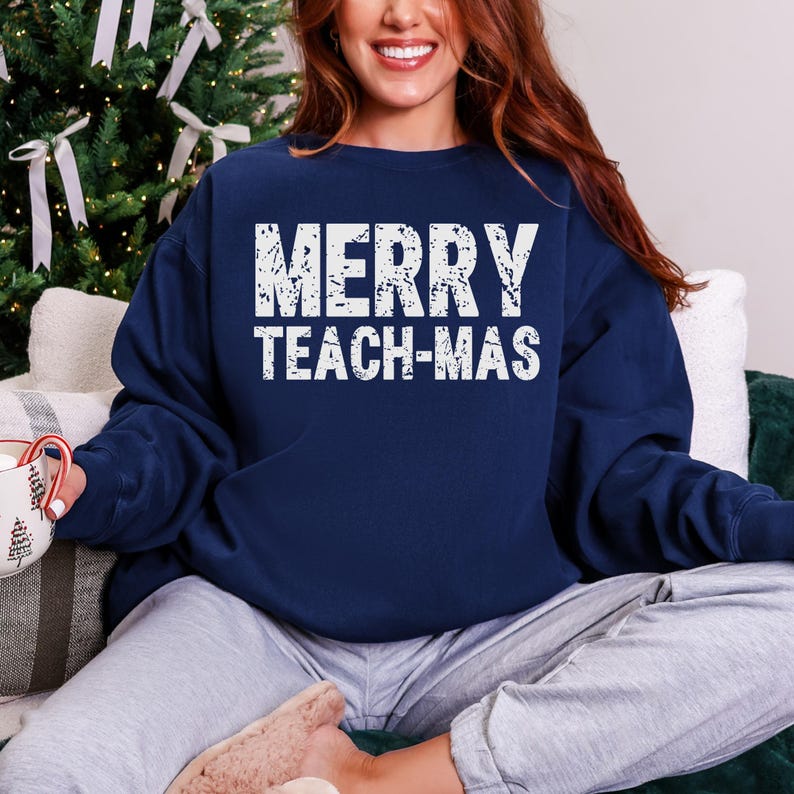 May include: Navy blue sweatshirt with the text "MERRY TEACH-MAS" in distressed white lettering. The sweatshirt is a crew neck style and is being worn by a person. The background includes a Christmas tree and a mug.