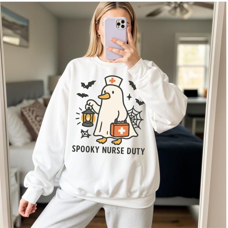 May include: White sweatshirt with a Halloween-themed graphic. The design features a ghost-like duck wearing a nurse's cap and carrying a lantern and medical bag. The text "SPOOKY NURSE DUTY" is printed below the graphic, with bats and a spiderweb completing the design.