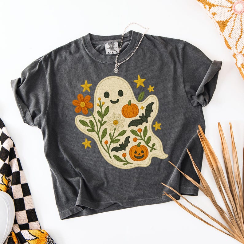 May include: Dark grey t-shirt with a cream-coloured ghost design. The ghost has a smiling face, surrounded by orange pumpkins, yellow stars, and green foliage. A silver necklace with a pendant rests on the shirt.