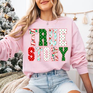May include: A light pink sweatshirt with the words "TRUE STORY" in a festive design. The letters are in red, green, and white, with patterns of polka dots and holiday-themed illustrations. The sweatshirt is a long-sleeved crew neck.
