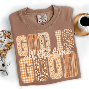 May include: A brown Comfort Colors t-shirt with the phrase "GOD IS all the time GOOD" in a mix of floral, plaid, and striped patterns. A cup of coffee sits on a wooden coaster to the right of the shirt.