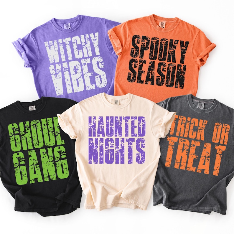 May include: Five colorful t-shirts with Halloween-themed phrases. The shirts are purple, orange, black, cream, and gray. The phrases include "Witchy Vibes," "Spooky Season," "Ghoul Gang," "Haunted Nights," and "Trick or Treat" in distressed fonts.